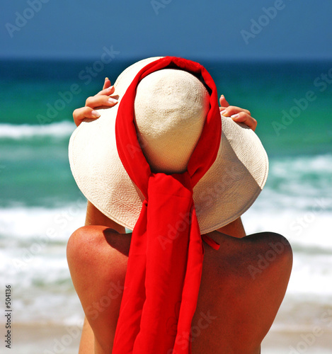 Photo: girl sitting on sandy beach in the sun adjusting hat. blue sky, girl sitting on sandy beach in the sun adjusting hat. blue sky,