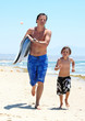 young father and son running along beach with surfboard