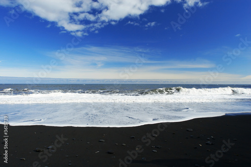 waves crashing on a sandy black beach , deep blue sky 