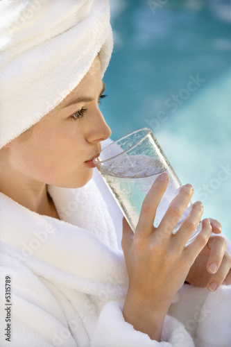 Photo: Woman wearing white terry robe drinking water next to pool. Woman wearing white terry robe drinking water next to pool.