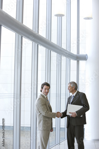 Photo: Two businessmen shaking hands in front of large glass windows Two businessmen shaking hands in front of large glass windows