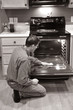 A man kneels on the floor in the kitchen and cleans the oven.