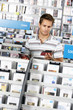 Photo: Young man choosing CD in record shop, standing in aisle behind rack, reading album sleeve (tilt) Young man choosing CD in record shop, standing in aisle behind rack, reading album sleeve (tilt)