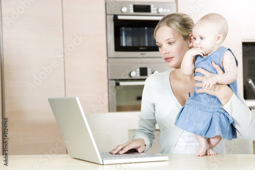 Photo: Mother using a laptop in the kitchen at home, holding baby daughter Mother using a laptop in the kitchen at home, holding baby daughter