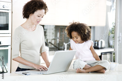 Photo: Mother using a laptop in the kitchen at home, while young daughter looks on Mother using a laptop in the kitchen at home, while young daughter looks on