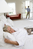 Photo: Mature woman lying in bed, smiling, portrait, man standing by window in background Mature woman lying in bed, smiling, portrait, man standing by window in background