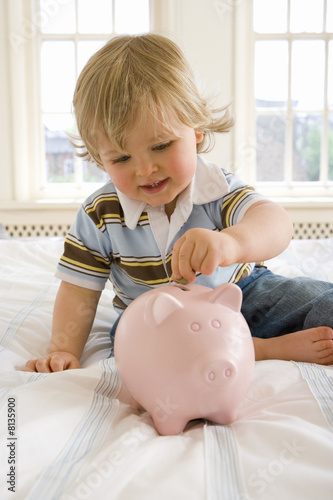 Foto: Baby boy (9-12 months) on bed putting coin in piggy bank, smiling, close-up Baby boy (9-12 months) on bed putting coin in piggy bank, smiling, close-up