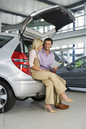 Photo: Couple looking at new silver hatchback in car showroom, sitting in open boot, man holding brochure, smiling, side view Couple looking at new silver hatchback in car showroom, sitting in open boot, man holding brochure, smiling, side view