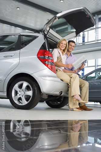 Photo: Couple looking at new silver hatchback in car showroom, sitting in open boot, man holding brochure, smiling, side view, portrait (surface level) Couple looking at new silver hatchback in car showroom, sitting in open boot, man holding brochure, smiling, side view, portrait (surface level)