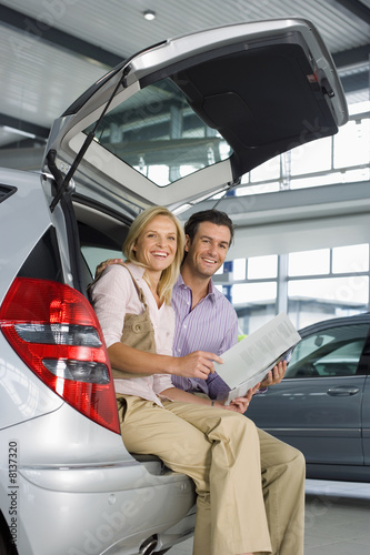 Photo: Couple looking at new silver hatchback in car showroom, sitting in open boot, man holding brochure, smiling, side view, portrait Couple looking at new silver hatchback in car showroom, sitting in open boot, man holding brochure, smiling, side view, portrait