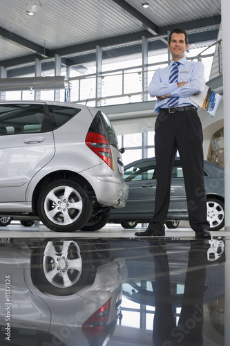 Photo: Car salesman standing near new cars in showroom, holding brochure, smiling, portrait (surface level) Car salesman standing near new cars in showroom, holding brochure, smiling, portrait (surface level)