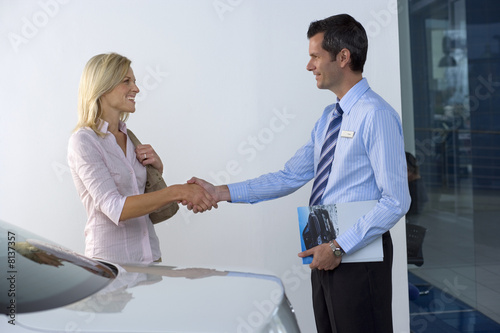 Photo: Car salesman shaking hands with female customer in showroom, man holding brochure, smiling, profile Car salesman shaking hands with female customer in showroom, man holding brochure, smiling, profile