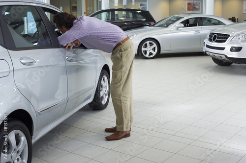 Photo: Man looking at new silver saloon car in showroom, leaning through open driver’s seat window, side view Man looking at new silver saloon car in showroom, leaning through open driver’s seat window, side view
