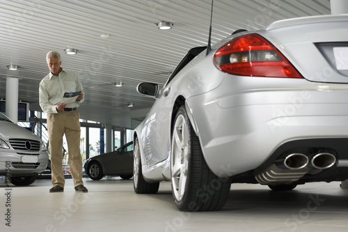 Photo: Senior man looking at new silver convertible car in large showroom, holding brochure (surface level) Senior man looking at new silver convertible car in large showroom, holding brochure (surface level)