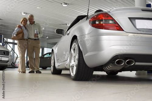 Photo: Senior couple looking at new silver convertible car in large showroom, man holding brochure, smiling (surface level) Senior couple looking at new silver convertible car in large showroom, man holding brochure, smiling (surface level)