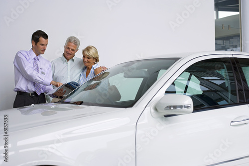 Photo: Car salesman showing senior couple new car in showroom, salesman pointing to brochure, smiling Car salesman showing senior couple new car in showroom, salesman pointing to brochure, smiling