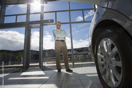 Photo: Senior man standing in car showroom, looking at new car (surface level, tilt, lens flare) Senior man standing in car showroom, looking at new car (surface level, tilt, lens flare)