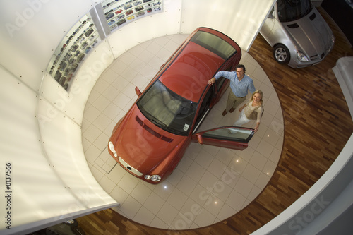 Photo: Couple looking at new red hatchback in car showroom, smiling, portrait, overhead view Couple looking at new red hatchback in car showroom, smiling, portrait, overhead view