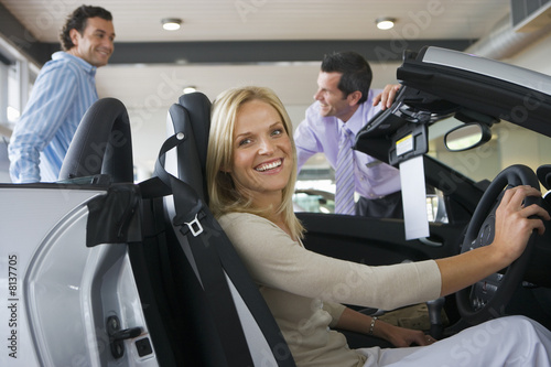 Photo: Car salesman showing couple new silver convertible in large car showroom, focus on woman sitting in driving seat in foreground, smiling, side view, portrait Car salesman showing couple new silver convertible in large car showroom, focus on woman sitting in driving seat in foreground, smiling, side view, portrait
