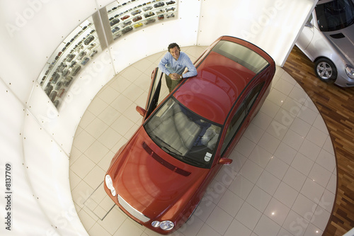 Photo: Man leaning against new red hatchback in large car showroom, smiling, portrait, overhead view Man leaning against new red hatchback in large car showroom, smiling, portrait, overhead view