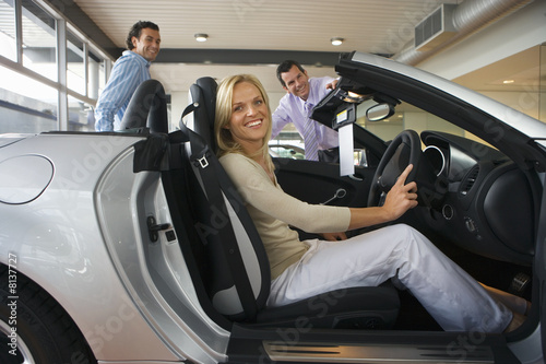 Photo: Car salesman showing couple new silver convertible in large car showroom, focus on woman sitting in driving seat in foreground, smiling, side view, portrait Car salesman showing couple new silver convertible in large car showroom, focus on woman sitting in driving seat in foreground, smiling, side view, portrait
