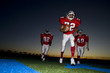 Photo: Three American football players, in red strips, running on pitch with ball at sunset (surface level) Three American football players, in red strips, running on pitch with ball at sunset (surface level)