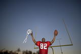 Photo: American football player, in red football strip, celebrating victory on pitch at sunset, arms up, holding ball and protective helmet in front of goal post, low angle view, portrait American football player, in red football strip, celebrating victory on pitch at sunset, arms up, holding ball and protective helmet in front of goal post, low angle view, portrait
