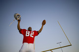 Photo: American football player, in red football strip, celebrating victory on pitch at sunset, arms up, holding ball and protective helmet in front of goal post, low angle view (tilt) American football player, in red football strip, celebrating victory on pitch at sunset, arms up, holding ball and protective helmet in front of goal post, low angle view (tilt)
