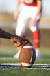 Photo: American football player attempting to kick field goal, focus on teammate holding ball vertically against pitch in foreground (surface level) American football player attempting to kick field goal, focus on teammate holding ball vertically against pitch in foreground (surface level)