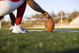 Photo: American football player attempting to kick field goal, teammate holding ball vertically against pitch (surface level) American football player attempting to kick field goal, teammate holding ball vertically against pitch (surface level)