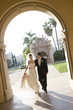 Photo: USA, California, San Diego, bride and groom walking arm in arm along pavement towards building arch, smiling, front view (tilt) USA, California, San Diego, bride and groom walking arm in arm along pavement towards building arch, smiling, front view (tilt)