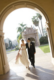 Photo: USA, California, San Diego, bride and groom walking arm in arm along pavement towards building arch, smiling, front view (tilt) USA, California, San Diego, bride and groom walking arm in arm along pavement towards building arch, smiling, front view (tilt)