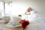 Photo: Breakfast tray with two coffee cups and single red rose on bed, focus on mature couple sitting upright in bed in background Breakfast tray with two coffee cups and single red rose on bed, focus on mature couple sitting upright in bed in background