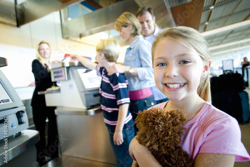 Photo: Family receiving ticket from female flight check-in attendant at airport check-in counter, focus on girl (7-9) holding soft toy in foreground, smiling, side view, portrait Family receiving ticket from female flight check-in attendant at airport check-in counter, focus on girl (7-9) holding soft toy in foreground, smiling, side view, portrait