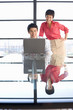 Photo: Young woman standing by young man sitting with laptop on glass table, smiling, window in background Young woman standing by young man sitting with laptop on glass table, smiling, window in background