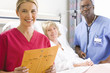 Photo: Male nurse with stethoscope by mature female patient in hospital bed, female nurse in foreground, smiling, portrait Male nurse with stethoscope by mature female patient in hospital bed, female nurse in foreground, smiling, portrait