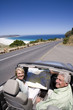 Photo: South Africa, Cape Town, senior couple with map parked on side of road by sea in convertible silver car, smiling, portrait, elevated view South Africa, Cape Town, senior couple with map parked on side of road by sea in convertible silver car, smiling, portrait, elevated view