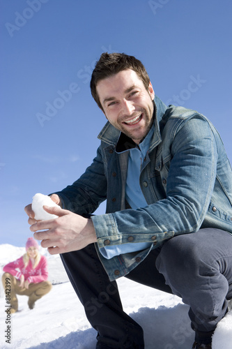 Photo: Young man crouching in snow field, holding snow ball, smiling, portrait, young woman in background Young man crouching in snow field, holding snow ball, smiling, portrait, young woman in background