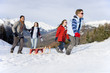 Photo: Boy and girl (7-9) pulling sled in snow field in front of parents, mountain range in background Boy and girl (7-9) pulling sled in snow field in front of parents, mountain range in background