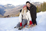 Photo: Young couple sitting on sled in snow field, smiling, portrait, mountain range in background Young couple sitting on sled in snow field, smiling, portrait, mountain range in background