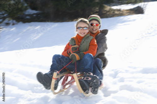 Photo: Boy and girl (7-9) riding sled down snow slope, wearing sunlgasses, smiling, low angle view Boy and girl (7-9) riding sled down snow slope, wearing sunlgasses, smiling, low angle view