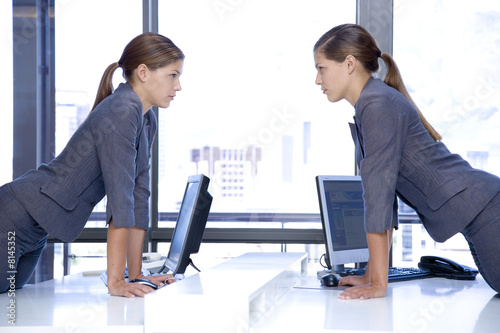 Photo: Identical female twins looking at each other over an office desk with computers Identical female twins looking at each other over an office desk with computers