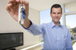 Photo: Male real estate agent holding up keys in living room, smiling, portrait (focus on keys) Male real estate agent holding up keys in living room, smiling, portrait (focus on keys)