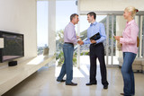 Photo: Male real estate agent standing with mature couple in living room, smiling and shaking hands with man Male real estate agent standing with mature couple in living room, smiling and shaking hands with man