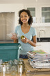 Photo: Young woman putting glass bottle and can into recycling bin in kitchen, smiling, portrait Young woman putting glass bottle and can into recycling bin in kitchen, smiling, portrait