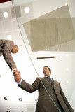 Photo: Two businessmen shaking hands by paperwork and pen on glass table, low angle view through glass Two businessmen shaking hands by paperwork and pen on glass table, low angle view through glass