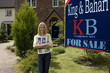 Young woman with magazine outside house with 'for sale' sign, smiling, portrait