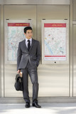 Photo: Businessman with briefcase and newspaper listening to MP3 player, by maps on wall Businessman with briefcase and newspaper listening to MP3 player, by maps on wall