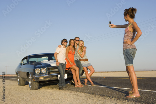 Photo: Young woman taking photograph of friends by car on desert road, low angle view Young woman taking photograph of friends by car on desert road, low angle view