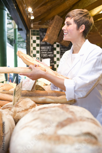 Photo: Female baker with baguettes in bakery, smiling, side view Female baker with baguettes in bakery, smiling, side view
